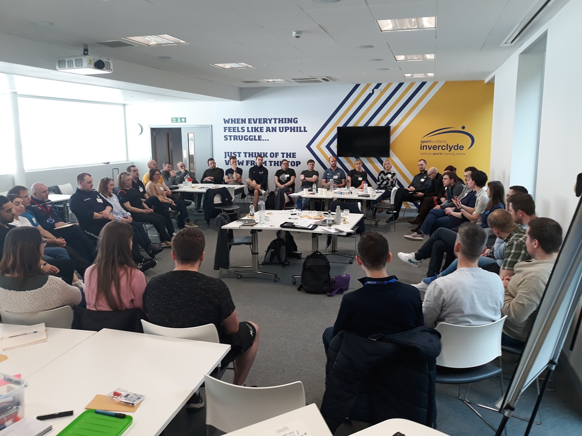 A group meeting, everyone is sitting around the outer edge of the room facing each other and listening to a person talk.