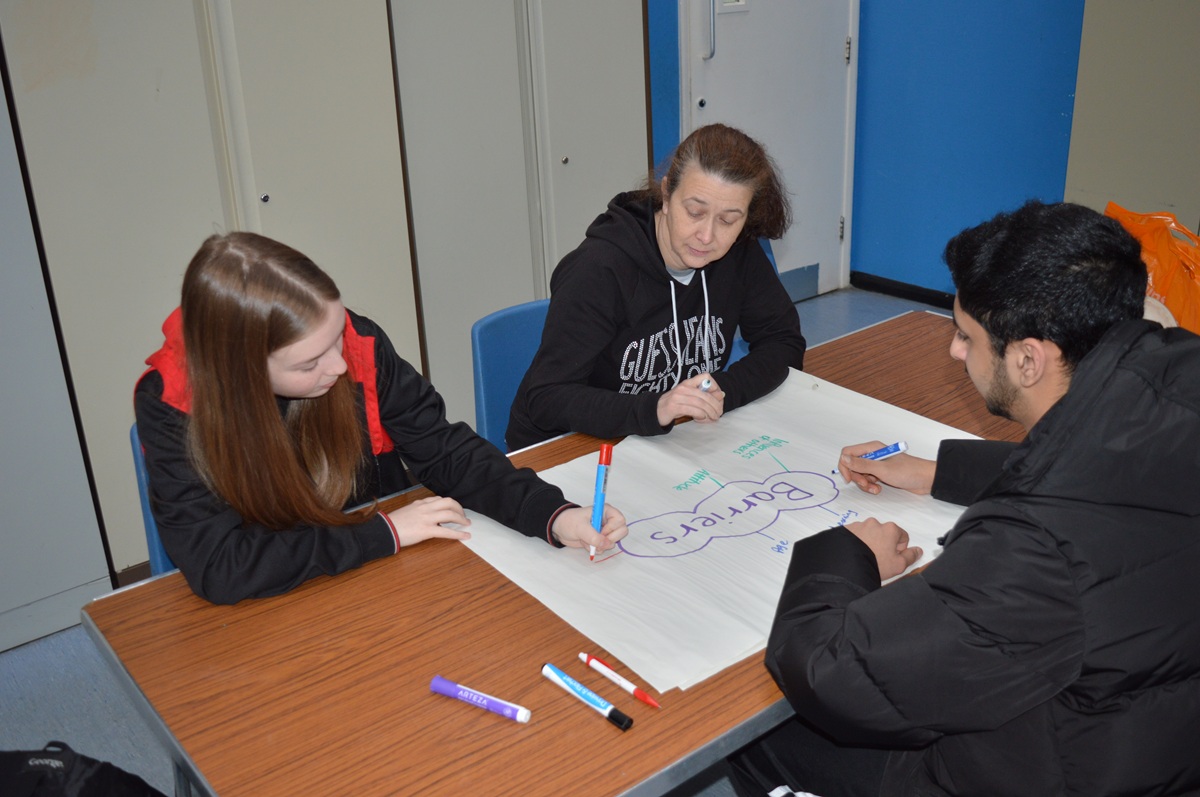 Three people sitting around an A1 sheet of paper adding words to a mind map that has barriers written in the middle. 