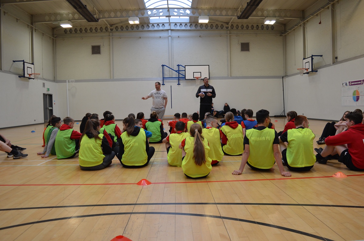 Two coaches talking to a group of students in a sports hall. The students are sitting in front of the two coaches. 