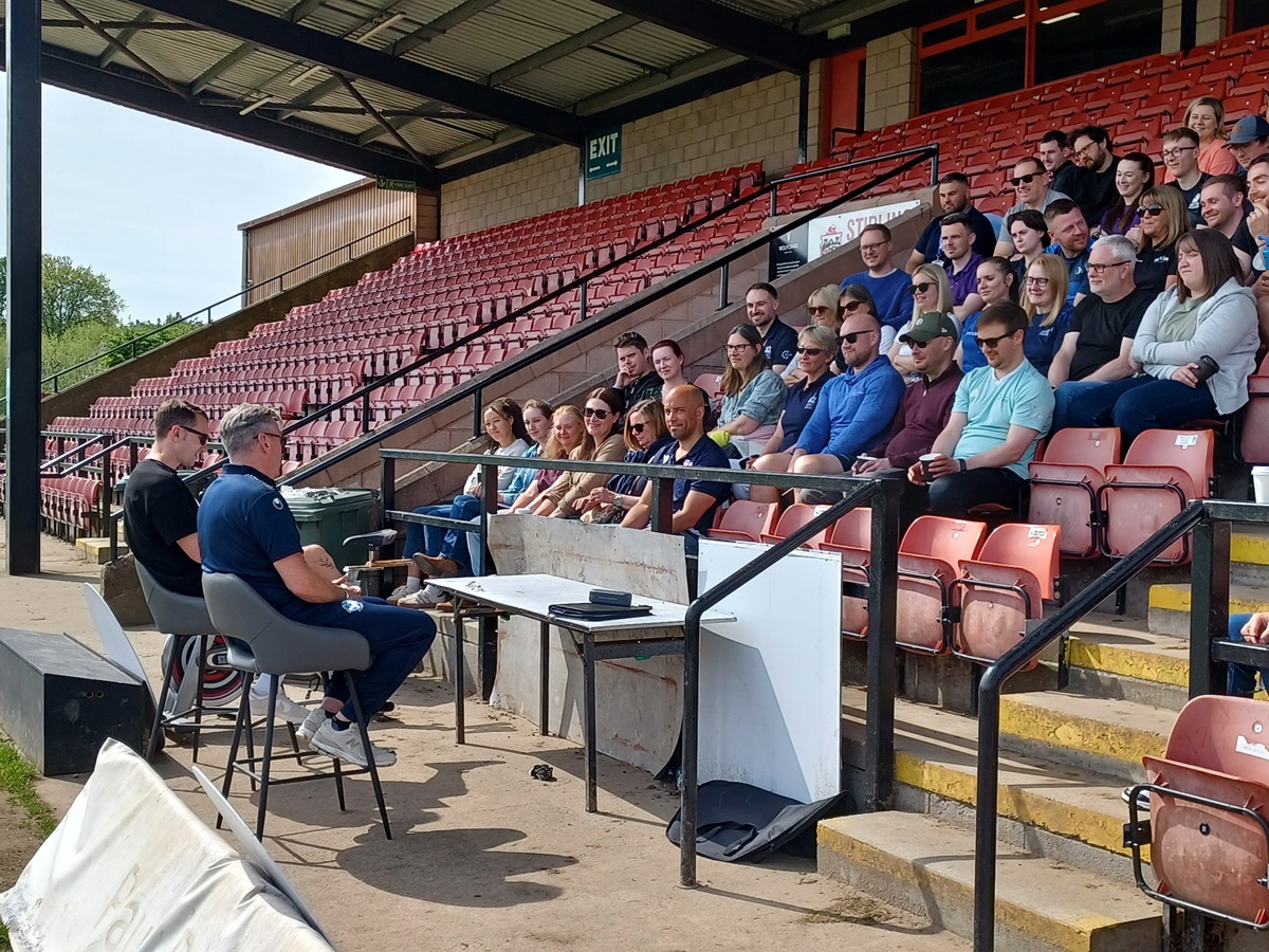 A meeting taking place in the stands of a sport venue, to people are sitting at the front talking to people in the stands. 